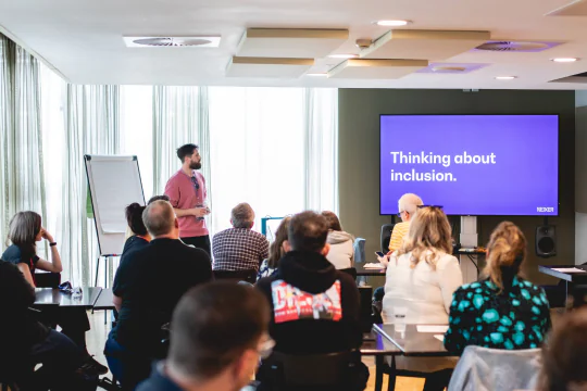 Danny Lancaster delivers a talk to a room of seated people. Onscreen is the text reading 'Thinking about inclusion'